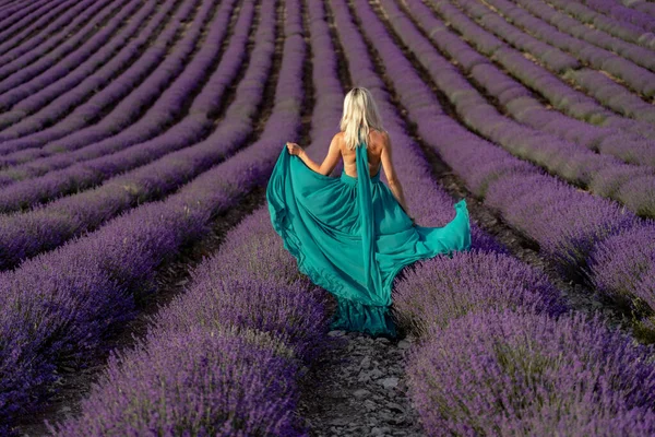 A woman with long blonde hair in a light green dress is walking through a lavender field alone waving a lavender dress. — Stock Photo, Image