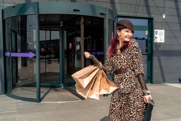 Szczęśliwa zakupoholiczka trzyma torby w pobliżu centrum handlowego. Kobieta w pobliżu sklepu jest zadowolona z zakupów, trzymając torby. Ubrana w sukienkę z lamparta. Koncepcja konsumenta. — Zdjęcie stockowe