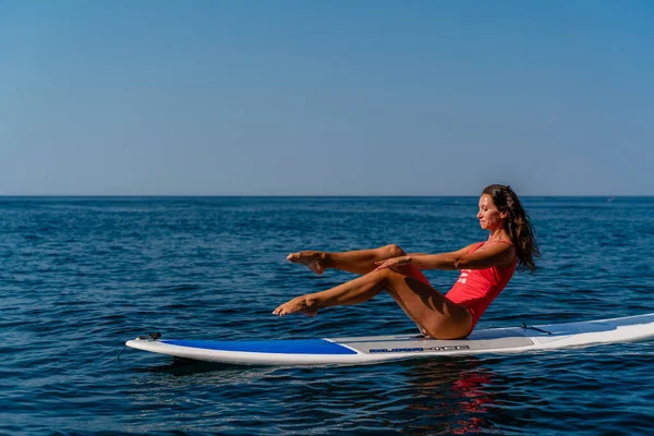 Спортивная девушка на доске для серфинга в море в солнечный летний день. В красном купальнике она сидит в шпагате на соке. Летние развлечения на Шторме у моря — стоковое фото