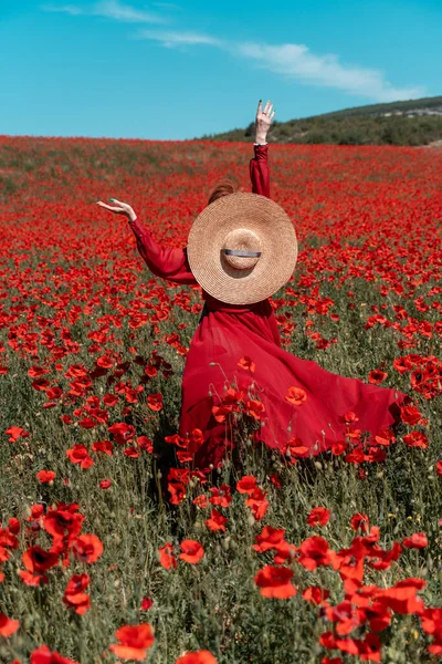 Jovem mulher fica de costas em um longo vestido vermelho e chapéu, posando em um grande campo de papoilas vermelhas — Fotografia de Stock