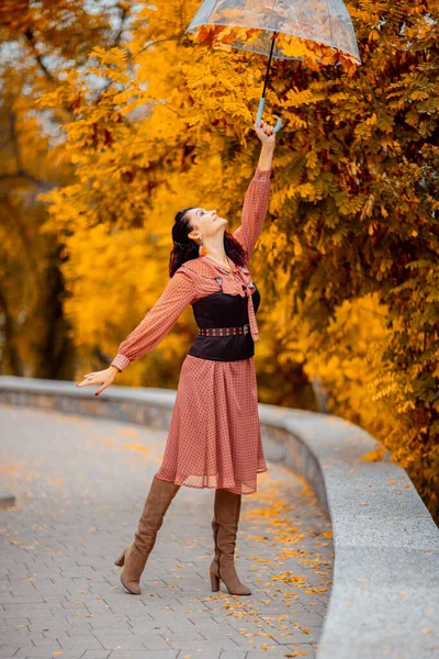 Hermosa chica en un vestido con un paraguas en el parque de otoño. Ella lo sostiene sobre su cabeza, las hojas de otoño caen de él —  Fotos de Stock