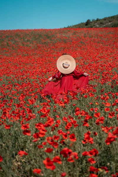 Jovem mulher fica de costas em um longo vestido vermelho e chapéu, posando em um grande campo de papoilas vermelhas — Fotografia de Stock