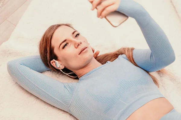 Портрет розслабленої жінки, що слухає музику з навушниками, що лежать на килимі вдома. Вона одягнена в синій трек-костюм . — стокове фото