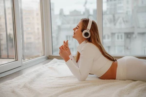 Retrato de vista lateral de una mujer relajada escuchando música con auriculares tumbados en la alfombra en casa. Ella está vestida con un chándal blanco. —  Fotos de Stock