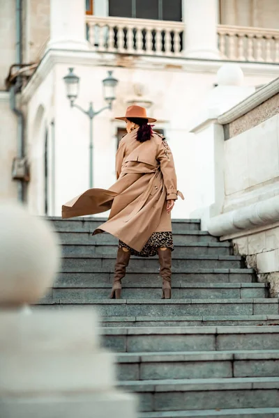 Odkryty portret mody młodej eleganckiej, modnej brunetki, modelki w stylowej czapce, chokerze i jasnym płaszczu przeciwdeszczowym, pozującej o zachodzie słońca w europejskim mieście. — Zdjęcie stockowe
