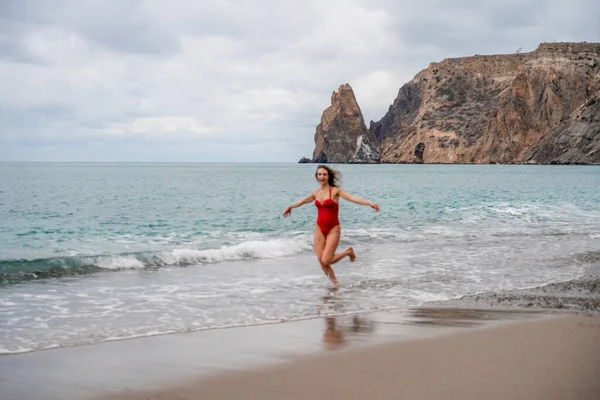 En vacker och sexig brunett i en röd baddräkt på en stenstrand, som löper längs stranden i vågornas skum — Stockfoto