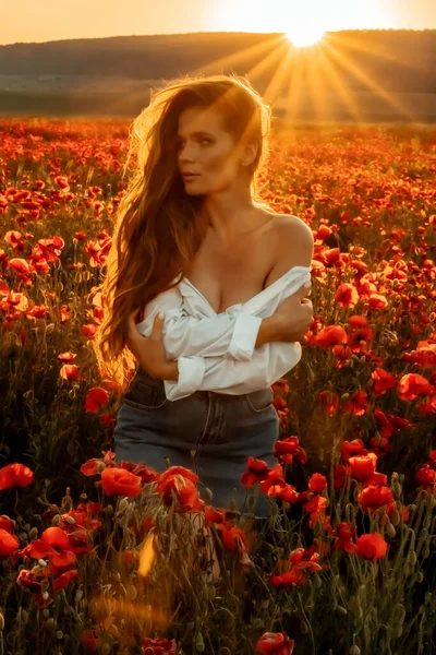 Bela jovem mulher de cabelos longos em um campo de papoula ao pôr do sol com cabelo voador. Vestido com uma camisa branca e saia jeans. — Fotografia de Stock