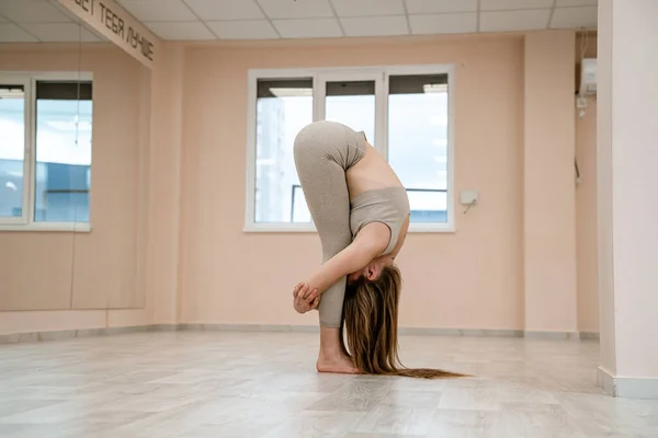 Wanita muda melakukan yoga di gym. Seorang gadis dengan kepang panjang dan dalam tracksuit krem berdiri di pose bangau di karpet merah muda. Seorang wanita melakukan padahstasana. — Stok Foto