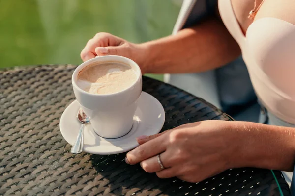 Primer plano horizontal de una joven sentada en un café sosteniendo una taza de café con leche. Retrato de estilo de vida interior de una chica irreconocible sosteniendo café latte bellamente decorado. — Foto de Stock