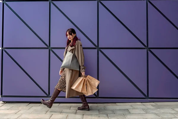 Szczęśliwa zakupoholiczka trzyma torby w pobliżu centrum handlowego. Kobieta w pobliżu sklepu jest zadowolona z zakupów, trzymając torby. Ubrana w sukienkę z lamparta. Koncepcja konsumenta. — Zdjęcie stockowe
