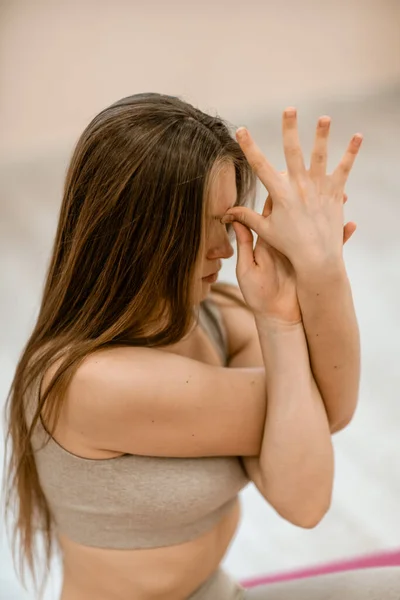 Mujer joven haciendo yoga en el gimnasio. Una chica con el pelo largo y un chándal beige se para en una pose de vaca sobre una alfombra rosa. Una mujer realiza Gomukhasana. —  Fotos de Stock