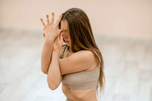 Mujer joven haciendo yoga en el gimnasio. Una chica con el pelo largo y un chándal beige se para en una pose de vaca sobre una alfombra rosa. Una mujer realiza Gomukhasana. — Foto de Stock