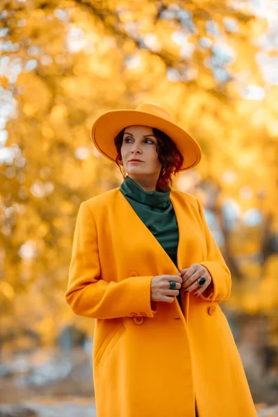 Hermosa mujer camina al aire libre en otoño. Lleva un abrigo amarillo, un sombrero amarillo y un vestido verde. Mujer joven disfrutando del clima otoñal. Contenido de otoño —  Fotos de Stock