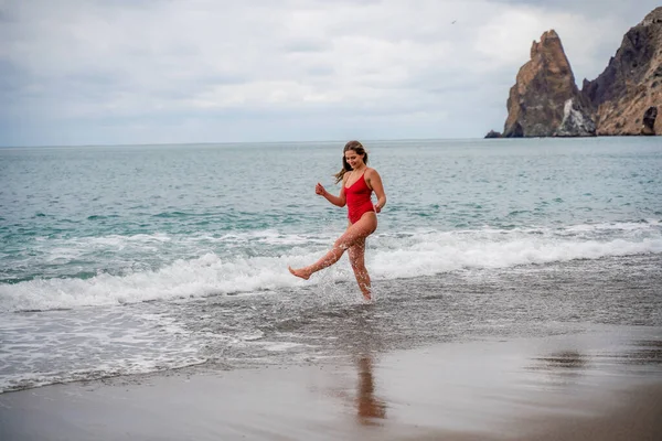 A beautiful and sexy brunette in a red swimsuit on a pebble beach, Running along the shore in the foam of the waves — Stock Photo, Image