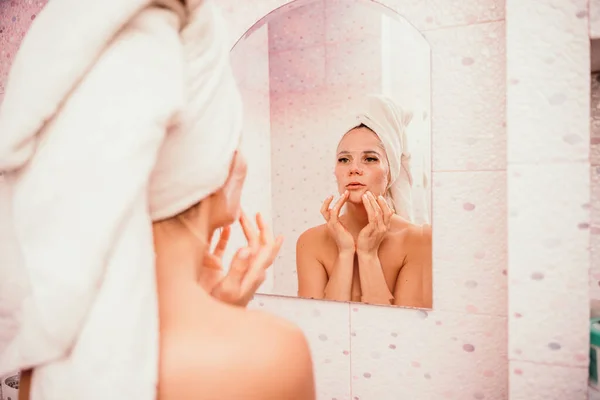 Mujer hermosa joven usando una máscara facial hidratante después de tomar un baño. Chica bastante atractiva en una toalla en la cabeza se para frente a un espejo en un baño en casa. Higiene diaria y cuidado de la piel — Foto de Stock