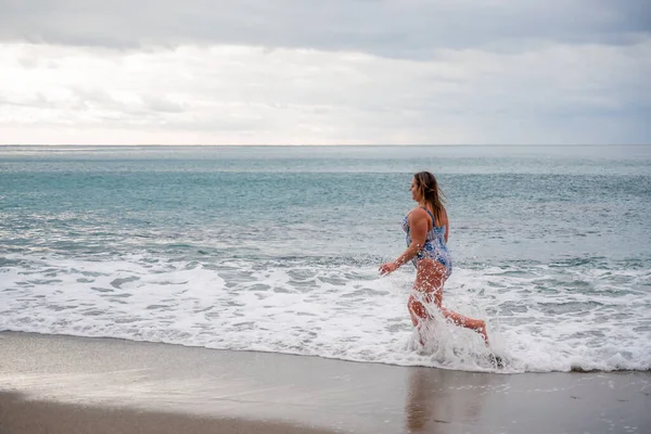 Una mujer gorda en traje de baño entra al agua durante el oleaje. Solo en la playa, cielo gris en las nubes, nadando en invierno. — Foto de Stock