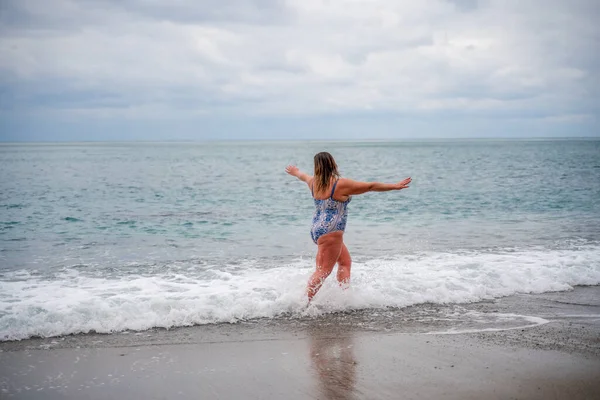 Eine dicke Frau im Badeanzug steigt während der Brandung ins Wasser. Allein am Strand, grauer Himmel in den Wolken, Schwimmen im Winter. — Stockfoto
