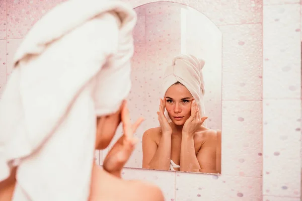 Jovem mulher bonita usando creme facial hidratante loção após o banho. Menina muito atraente em uma toalha na cabeça fica na frente de um espelho em uma casa de banho em casa. Higiene diária e cuidados com a pele — Fotografia de Stock