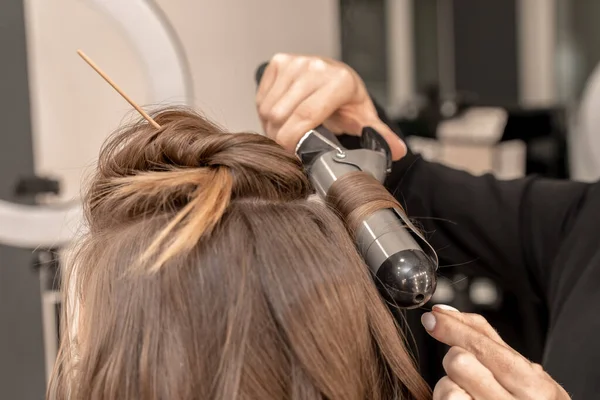 미용사는 여자를 준비시키고 , 컬링 철을 사용하여 곱슬머리로 머리를 만든다. 갈색의 짙은 갈색의 긴 털. 자기 관리 개념. — 스톡 사진