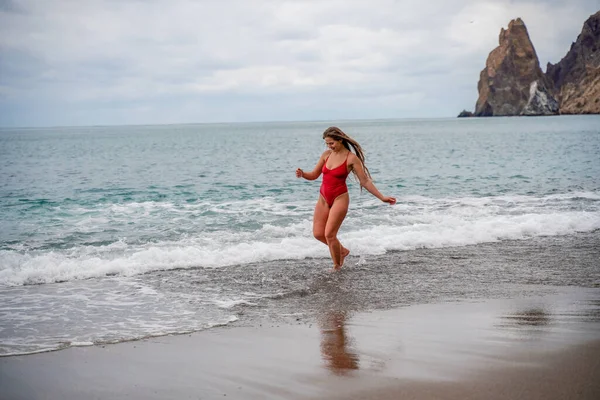 A beautiful and sexy brunette in a red swimsuit on a pebble beach, Running along the shore in the foam of the waves — Stock Photo, Image