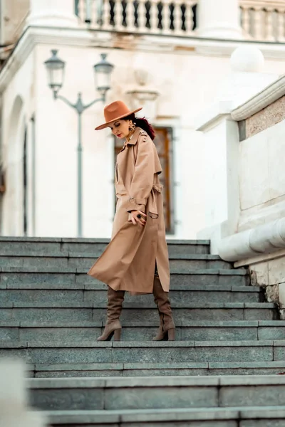Retrato de moda al aire libre de la joven mujer morena elegante de moda, modelo en sombrero elegante, gargantilla y impermeable ligero posando al atardecer en la ciudad europea. — Foto de Stock