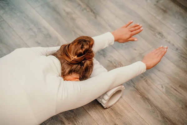 흰옷을 입은 성인 운동 선수인 여자 가 바닥에서 파시아 운동을 하고 있다 - 코카서스 여자가 안마 폼 롤러 - 등의 긴장을 완화하고 근육 통증을 완화하는 도구 - 를 사용하고 있다 — 스톡 사진
