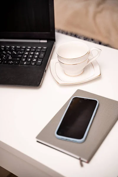 White desktop with laptop, coffee mug, phone and notebook for remote work or study. Office computer workplace in home interior.