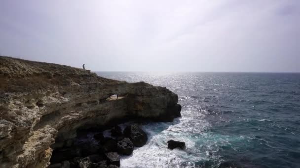 青いジャケットの女性は 海を見下ろす崖の上を歩き 嵐のような海を見ています 女の子旅行者は休息し 夢を見 自然を楽しむ 平和と穏やかな風景 風の強い天気 — ストック動画