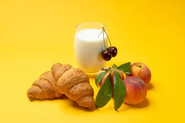 Frehch kruvasan, bir bardak süt, sarı arka planda taze şeftali ve kiraz. Yaz kahvaltısı