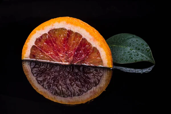 Slice of red sicilian orange fruit  and green leaf on black reflective background. Fresh, ripe bloody orange wallpaper