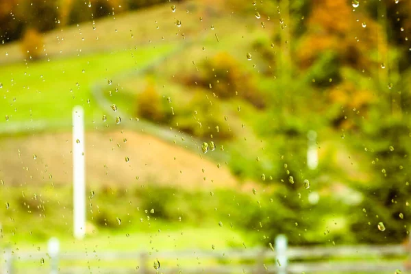 Rain drops on the window with green landscape on the background. Autumn weather in the village. Rainy day out of the window