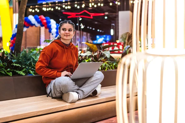 Bankta oturan genç bir kadın dizüstü bilgisayarıyla alışveriş merkezinin önünde bir lambayla bacak bacak bacak üstüne atmış. — Stok fotoğraf
