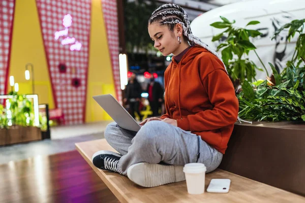 Mujer joven usando su portátil sentado en el banco con las piernas cruzadas en un centro comercial — Foto de Stock