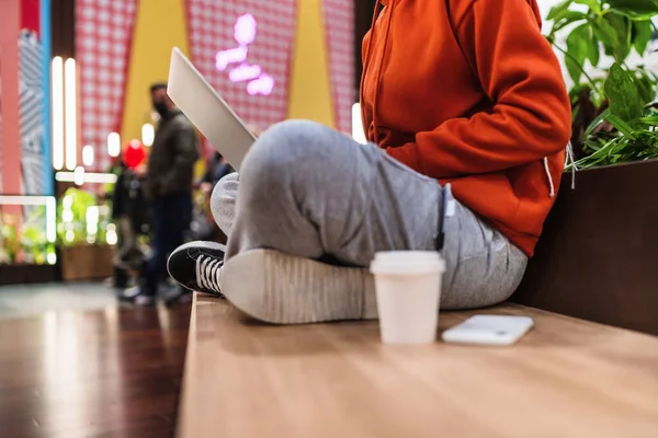 Vista recortada de una joven irreconocible sentada en el banco con las piernas cruzadas usando su computadora portátil en un centro comercial — Foto de Stock