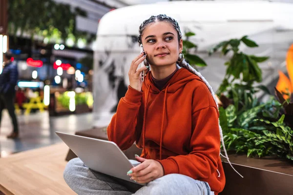 Una joven sentada en el banco con su portátil hablando con su teléfono en un centro comercial — Foto de Stock