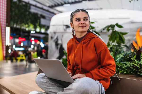 Mujer joven sentada en un banco sonriendo y mirando hacia otro lado mientras usa su computadora portátil en un centro comercial — Foto de Stock