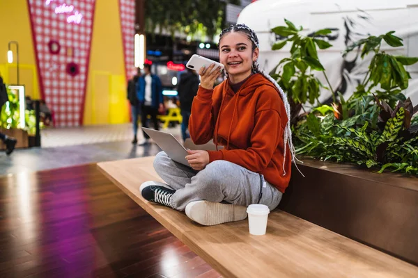 Eine junge Frau sitzt mit ihrem Laptop auf einer Bank und nimmt eine Audiobotschaft in einem Einkaufszentrum auf — Stockfoto
