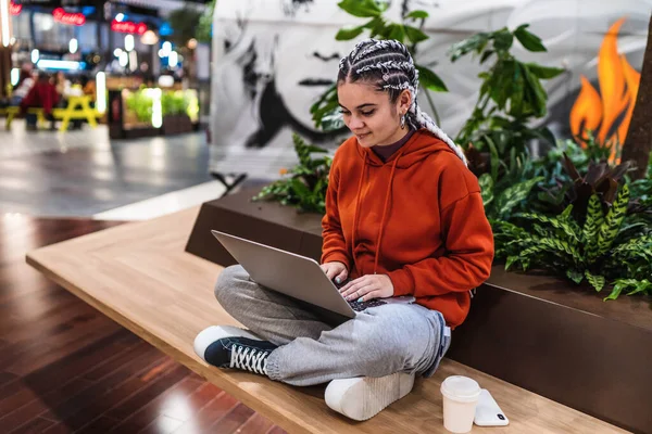 Vista de ángulo alto de una mujer joven usando su computadora portátil sentada en el banco con las piernas cruzadas en un centro comercial — Foto de Stock