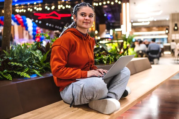 Una joven feliz sentada en el banco con las piernas cruzadas usando su portátil en un centro comercial — Foto de Stock
