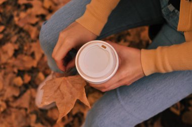 Paper glass of coffee in hand on the orange autumn leaves background . High quality photo