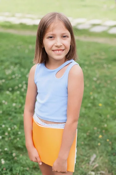 Portrét půvabné holčičky ve věku 8-10 let v tričku a šortkách se žlutou knihou v rukou, na letní zelené louce, filtrovaný obraz. vzdělávání — Stock fotografie