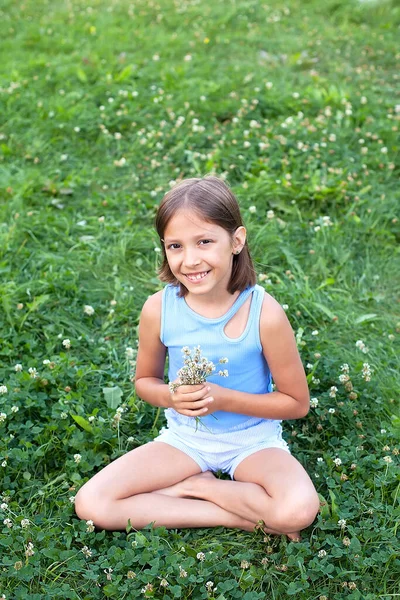 Uma menina adolescente em uma camiseta e shorts em uma clareira com grama verde gosta de verão em um dia ensolarado, olha para a câmera e sorri. Feliz infância. Férias de verão — Fotografia de Stock