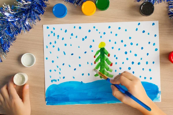 Un niño pinta un árbol de Navidad y nieve en un pedazo de papel. Dibujo de Año Nuevo para niños. Gouache y cepillos. Desarrollo infantil —  Fotos de Stock