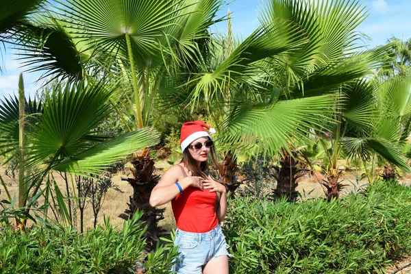 サンタクロースの帽子の女の子と緑のヤシの木の背景に赤いTシャツ 暖かい国での新年とクリスマスの休日の概念 — ストック写真