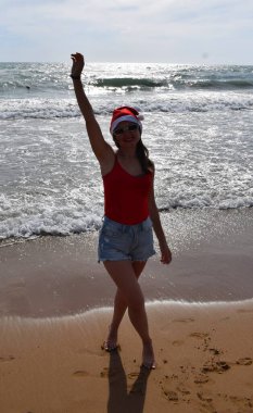 Noel Baba şapkalı bir kız ve akşam denizin arka planında kırmızı tişörtlü bir kız. Sıcak ülkelerde yeni yıl ve Noel tatili kavramı