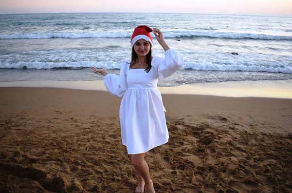 夜の海を背景にサンタクロースの帽子と白いドレスの女の子 暖かい国での新年とクリスマスの休日の概念 — ストック写真