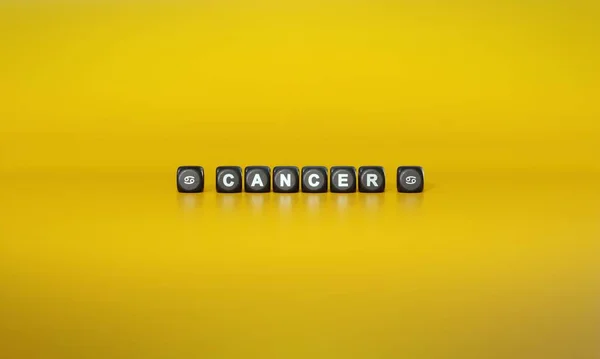Word Cancer and zodiac sign symbols spelled out in white text on dark wooden blocks against plain yellow background