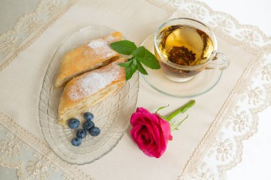 There are two slices of apple pie, blueberries and mint leaves on the plate, a cup of tea next to it, and the table is decorated with a live rose.