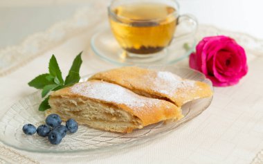 There are two slices of apple pie, blueberries and mint leaves on the plate, a cup of tea next to it, and the table is decorated with a live rose.