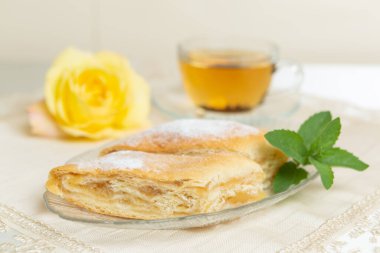 There are two slices of apple pie, blueberries and mint leaves on the plate, a cup of tea next to it, and the table is decorated with a live rose.